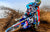 Motocross racer hitting a berm with loose dirt while on the throttle causing dirt to spray behind him as they ride through the turn? The rider has risk racing grips installed on their bike and are wearing risk racing motocross gear.
