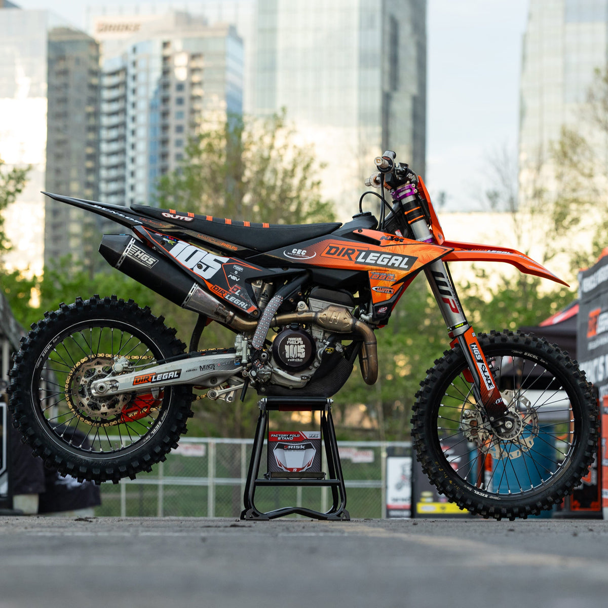 Moto de cross naranja y negra en un Risk Racing F.F.S. Stand con paisaje urbano en el fondo.