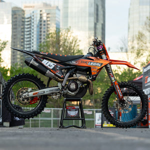 Moto de cross naranja y negra en un Risk Racing F.F.S. Stand con paisaje urbano en el fondo.